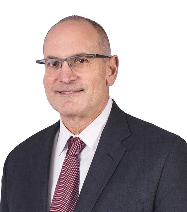 A photo of Ron Klemencic, wearing a black suit, a red tie, and glasses.